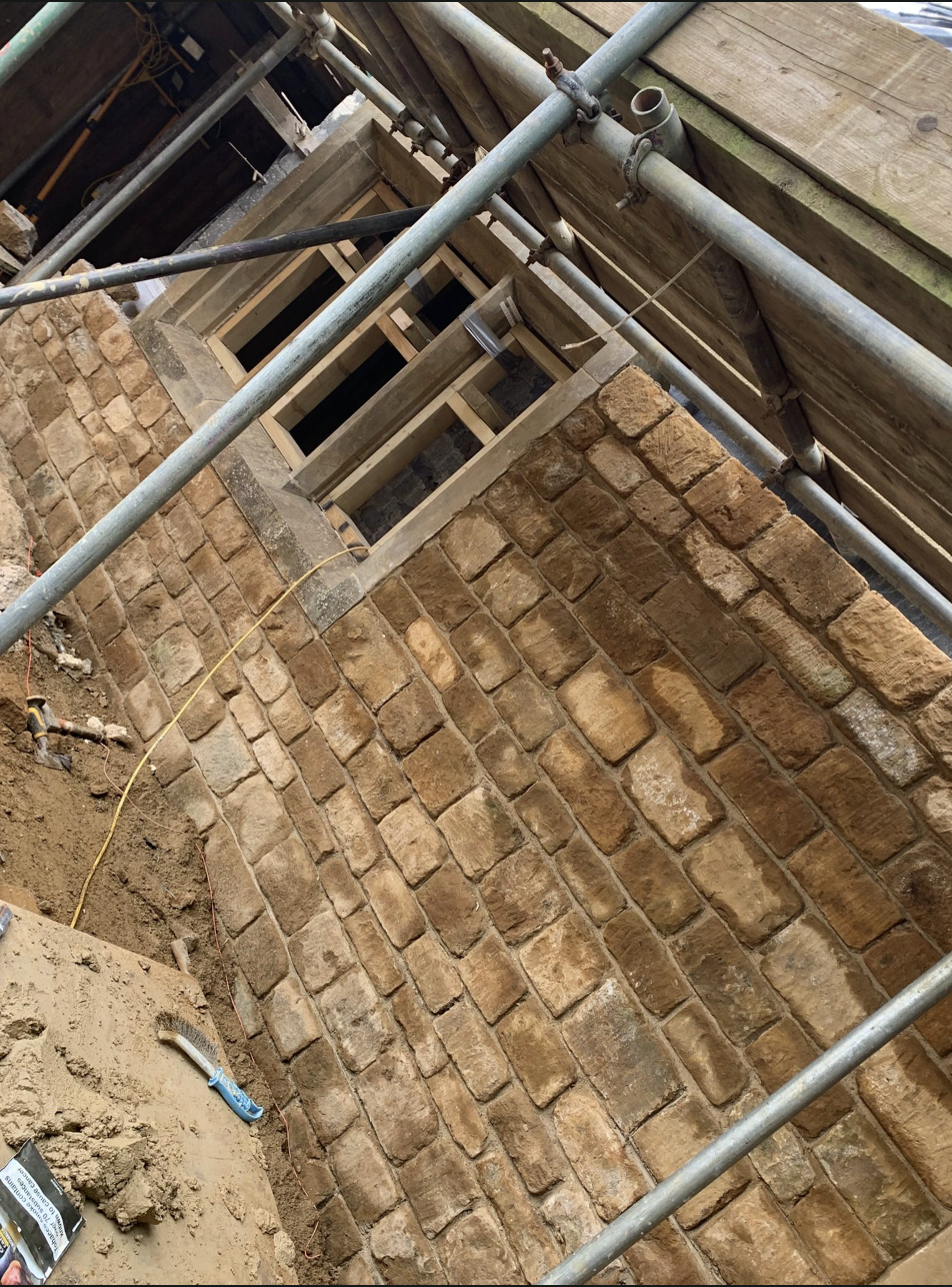 Cotswold stone wall mid-build, scaffolded, with dressed stones laid in fresh lime mortar